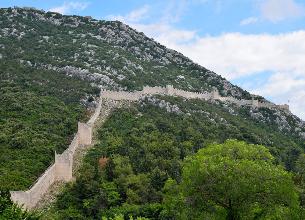 ...Im 14. Jahrhundert wurde mit dem Bau der Befestigungsanlagen begonnen. Es entstanden 3 Kastelle, die durch Mauern miteinander verbunden waren. Über 40 Türme waren Teil der ca. 5 km langen Mauer, von der bis heute noch Teile stehen. So war der Zugang zur Halbinsel Pelješac vollständig kontrollierbar. Die Gesamtanlage wurde 1506 fertig und wird als die längste Festungsmauer in Europa bezeichnet...